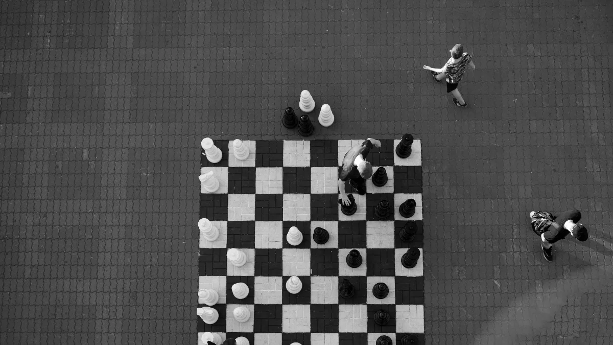 Top-down view of a giant outdoor chessboard with a person making a move, while others walk past. The image symbolizes strategic thinking and seeing the bigger picture